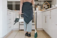 a woman stand in kitchen with broom and cleaning solutions in hands