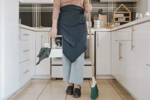 a woman stand in kitchen with broom and cleaning solutions in hands