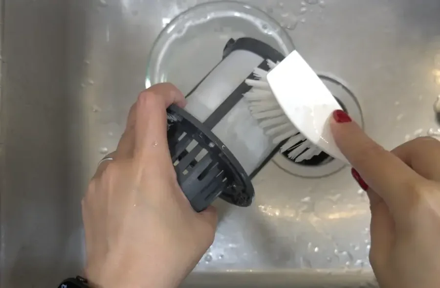Person scrubbing a removable dishwasher filter under the sink with a cleaning brush.