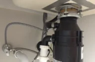 Black garbage disposal unit connected to white PVC plumbing and water lines under a kitchen sink