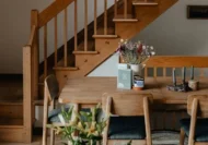 A cozy dining area featuring a natural mango wood table and chairs, accented with dried flowers and modern table décor, set against a wooden staircase backdrop